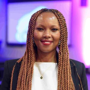 Winnie Karanu smiling in front of a screen with purple-blue lighting, wearing a black blazer and gold necklace—Microsoft’s AI Skills Director at a professional event.
