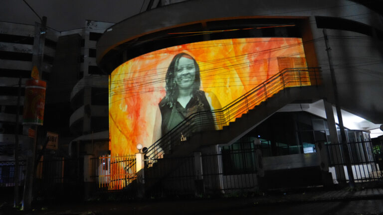 A curved building facade in Nairobi at night displays a large projected portrait of a smiling Kenyan woman against a vivid amber and red painted background, part of the Jute Portraits installation.