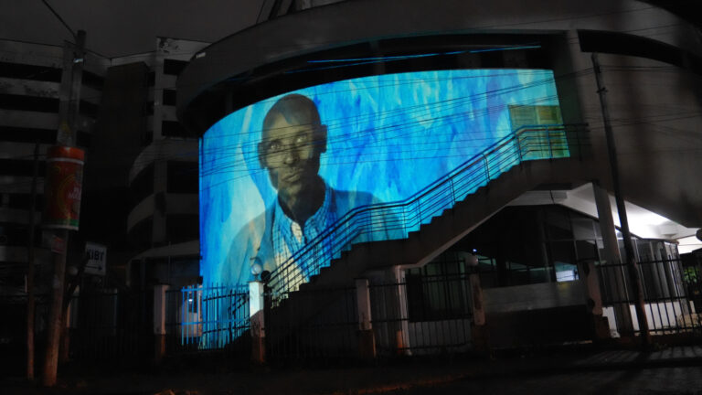 A curved building facade in Nairobi at night displays a projected portrait of a Kenyan man wearing glasses against a deep blue painted background, part of Franz Cerami's Jute Portraits public art installation.