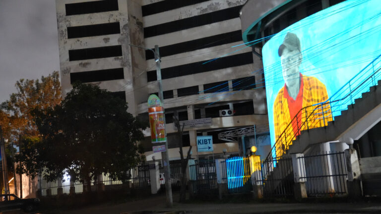 A large outdoor LED screen on a curved building facade in Nairobi displays a digitally painted portrait of a Kenyan coffee worker wearing a cap and checked shirt, projected in teal and aqua tones at night.