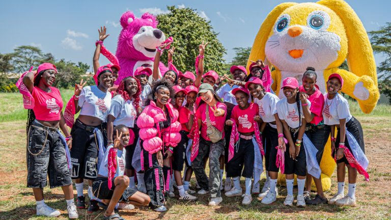 Svetlana Polikarpova, Director of SKY Girls Kenya, poses with a group of teenage girls in pink Peace Over Pressure T-shirts and caps at SKY FEST 2026 in Nairobi, flanked by two oversized mascots — a pink bear and a yellow rabbit — under a bright blue sky.