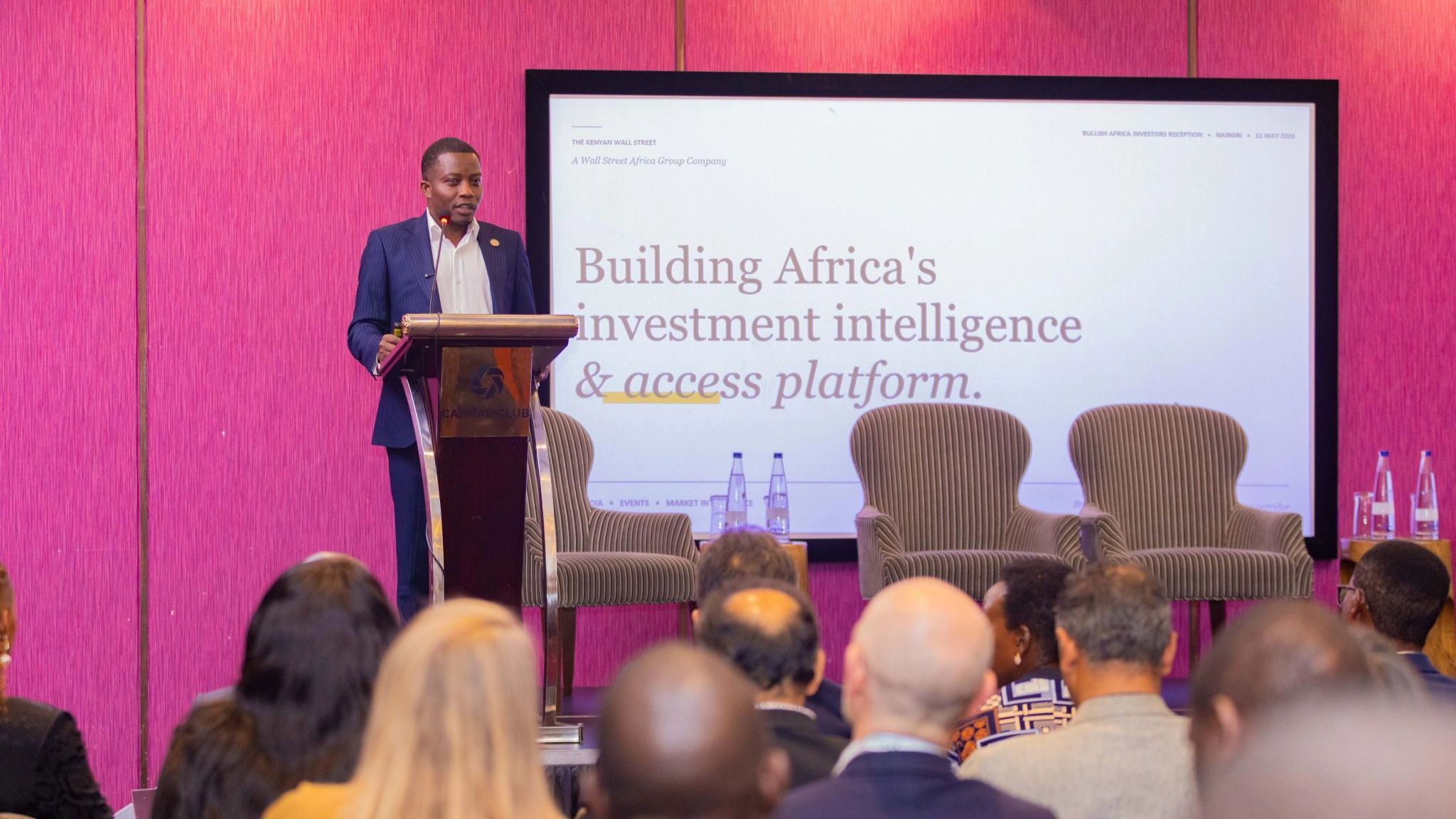 A man in a navy pinstripe suit speaks at a podium in front of a pink-walled conference room, with a screen behind him reading "Building Africa's investment intelligence & access platform."