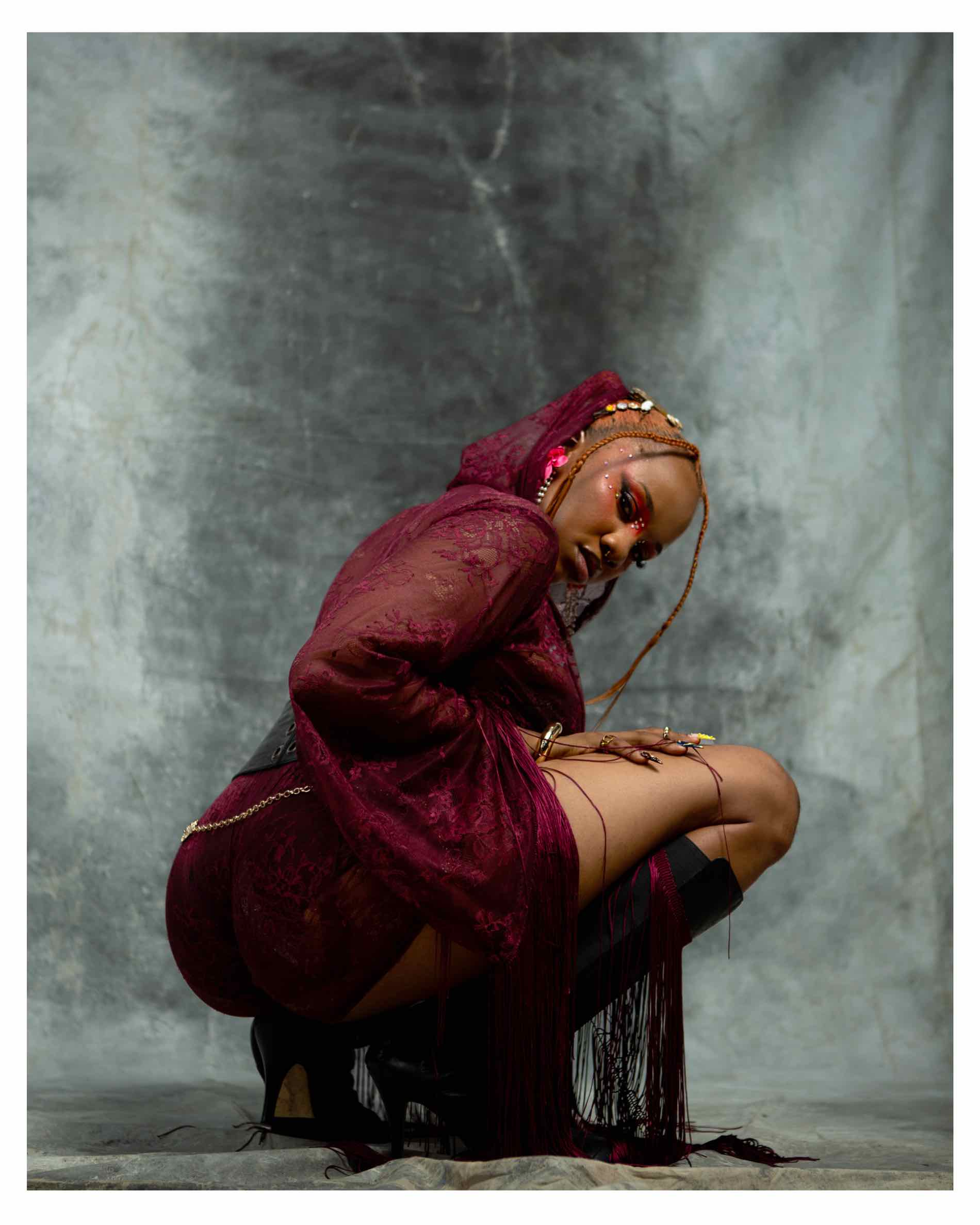 AfroReekAH crouched in a burgundy lace outfit against a grey backdrop for the REEPLAY campaign