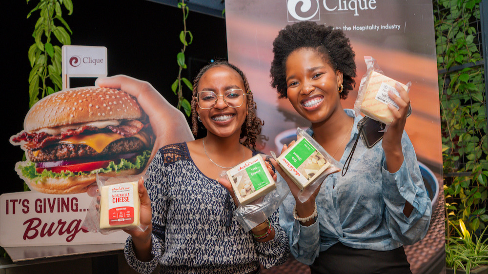 Two women hold packs of Cheese Love mozzarella and feta cheese in front of a Clique branded Burger Week display.