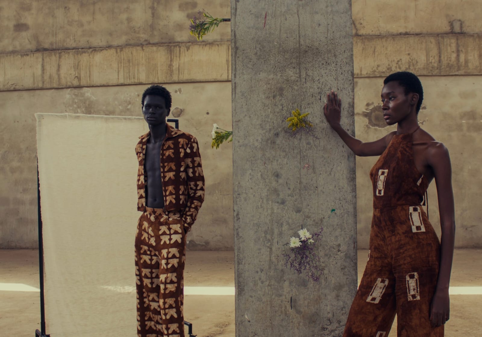Two models stand beside a concrete column, one in a brown printed jacket and trousers, the other in a halter neck jumpsuit