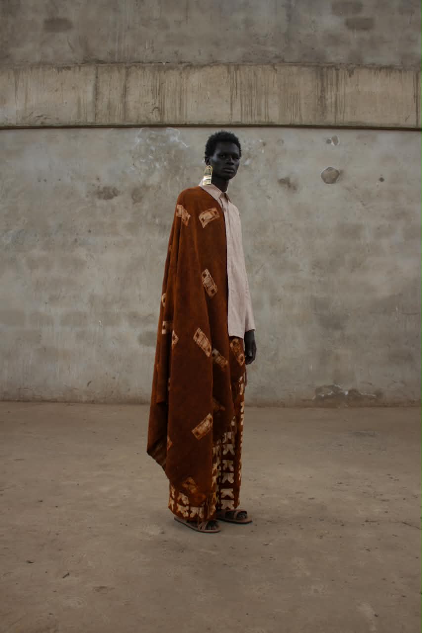 Model in a cream shirt wears a rust coloured cloak draped over the shoulder, with printed trousers