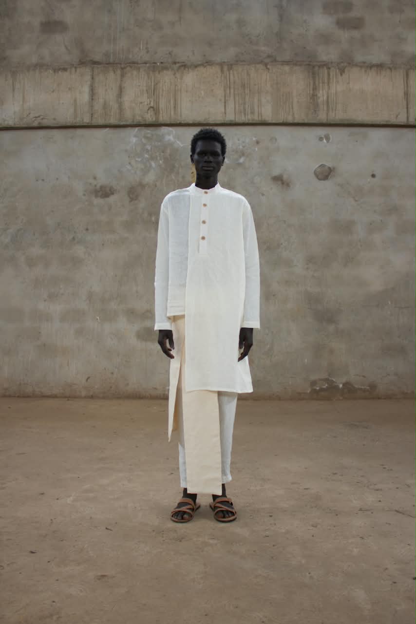 Model in a long white tunic with a stand collar and three buttons over white trousers with a side panel