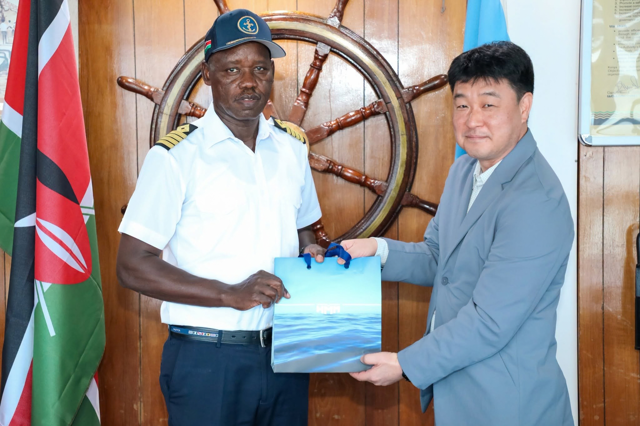 Man in a captain's uniform and man in a grey blazer hold an HMM gift bag before a ship's wheel and a Kenyan flag.