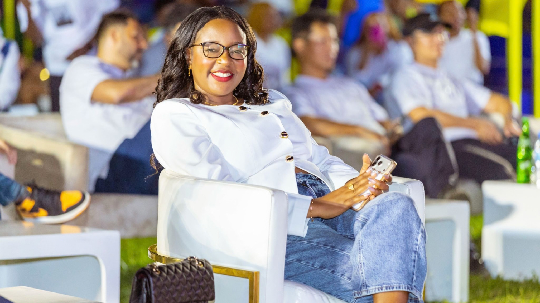 A woman in glasses and a white jacket sits in a white armchair at an outdoor evening event, holding a phone, with an audience behind her.