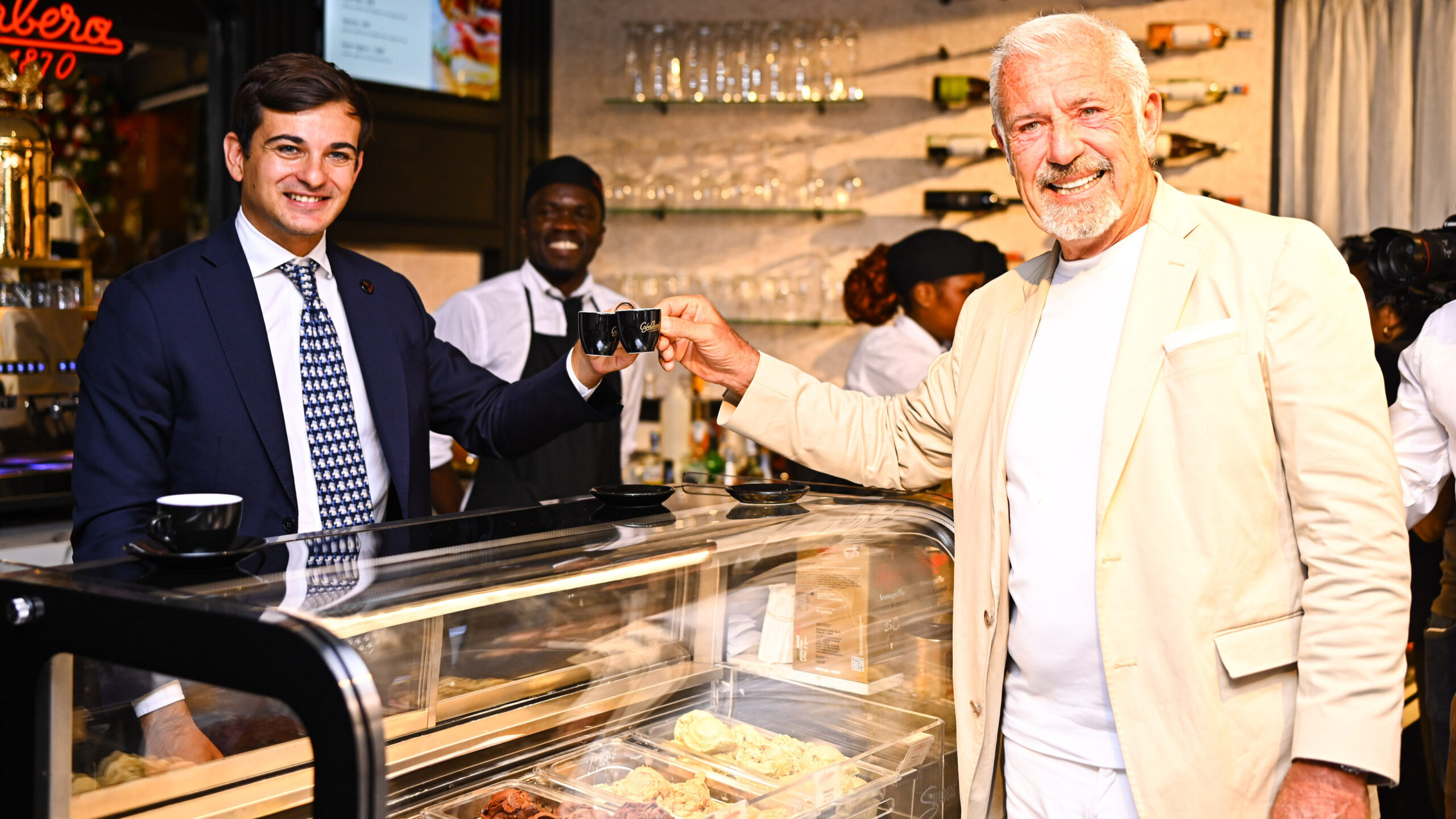 Two men touch espresso cups behind a gelato counter, with staff working in the background.