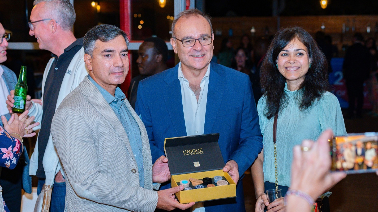 Three guests pose at an evening event, one holding an open gold box labelled "Unique and Repeatable" that contains small round items. A guest behind them holds a bottle, and a hand in the foreground holds up a phone to take a photo.