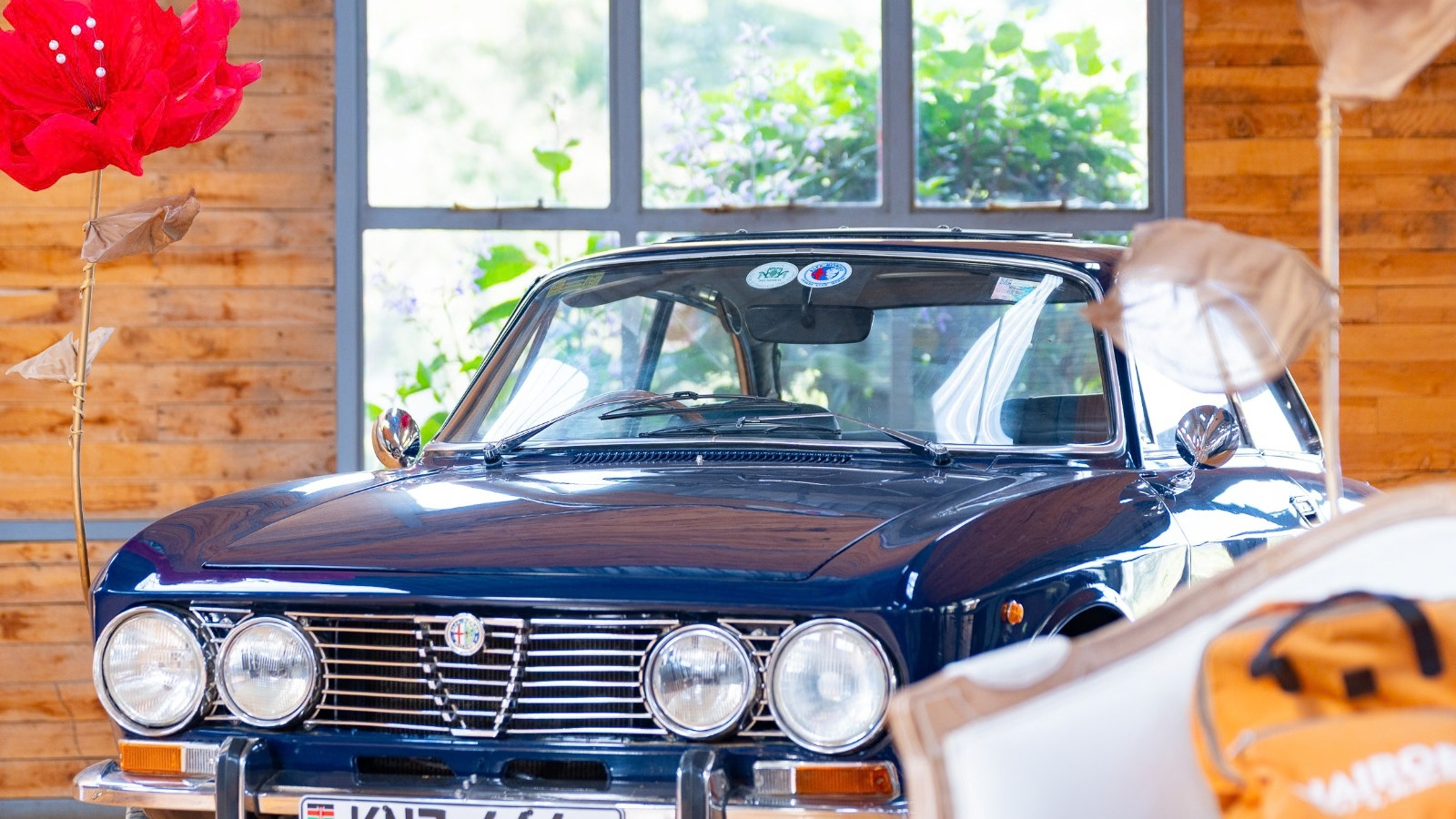 Front view of a dark blue vintage Alfa Romeo coupe with four round headlamps, a chrome shield grille and a Kenyan number plate. The car stands indoors against a timber wall and a window onto greenery, framed by large red paper flowers.