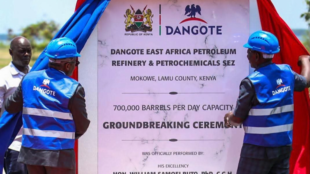 Two men in blue hard hats and blue Dangote safety vests pull back red and blue drapes from a marble plaque. The plaque names the Dangote East Africa Petroleum Refinery & Petrochemicals SEZ, Mokowe, Lamu County, 700,000 barrels per day capacity, and a groundbreaking ceremony on 30 September 2026.