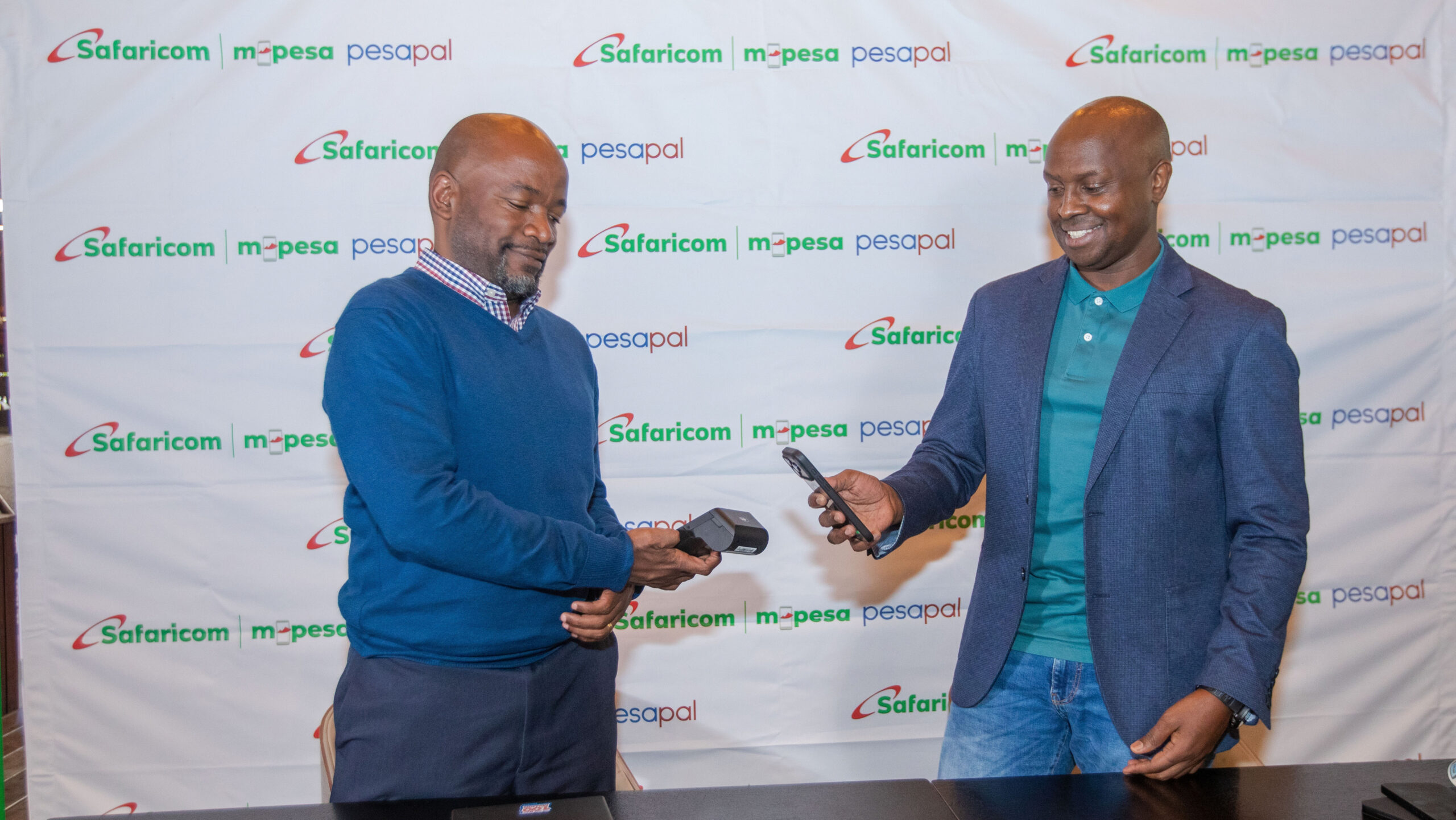Two men stand at a table in front of a Safaricom, M-PESA and Pesapal backdrop. The man on the left holds a point of sale device and the man on the right holds a smartphone near it to show a tap payment.