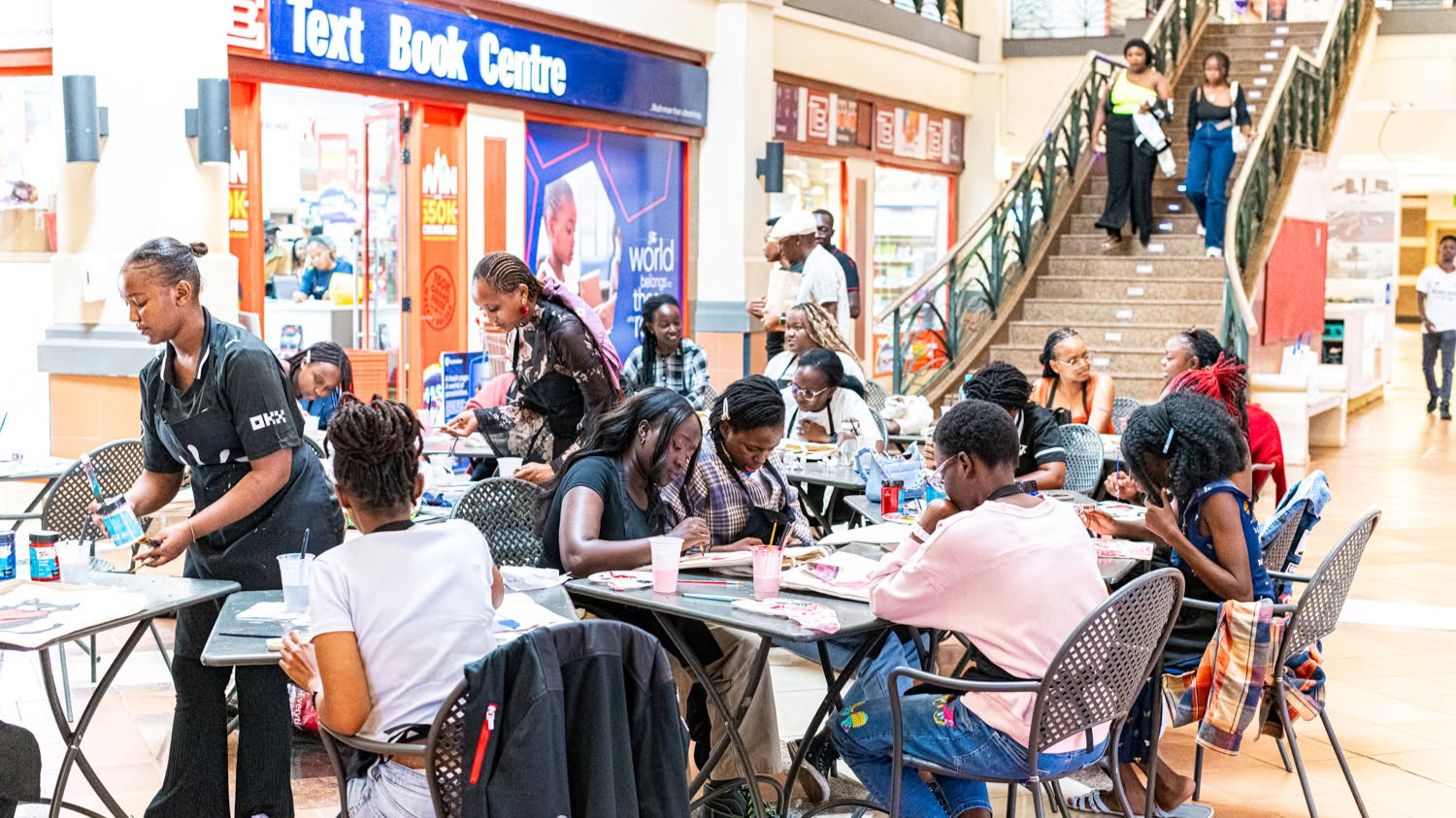 Participants seated at tables in the Galleria Shopping Mall corridor, painting custom designs on canvas tote bags outside Text Book Centre.