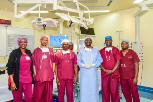 Dr. Bob Achila, a Consultant Urogynaecologist at Aga Khan University Hospital, is pictured third from the left in a group photo with fellow doctors, nurses, theater assistants, and technologists. The group is gathered in a surgical theater, likely after successfully performing a vNOTES surgical procedure.