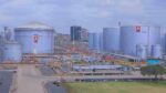 Industrial facility with large cylindrical KPC tanks and urban skyline. These tanks are interconnected by a dense network of pipes, staircases, and access platforms. In the backdrop, a mix of commercial and residential buildings suggests the site's integration within an urban or peri-urban environment. The layout implies petroleum or chemical storage operations at scale, emphasizing infrastructure complexity and proximity to strategic distribution corridors.