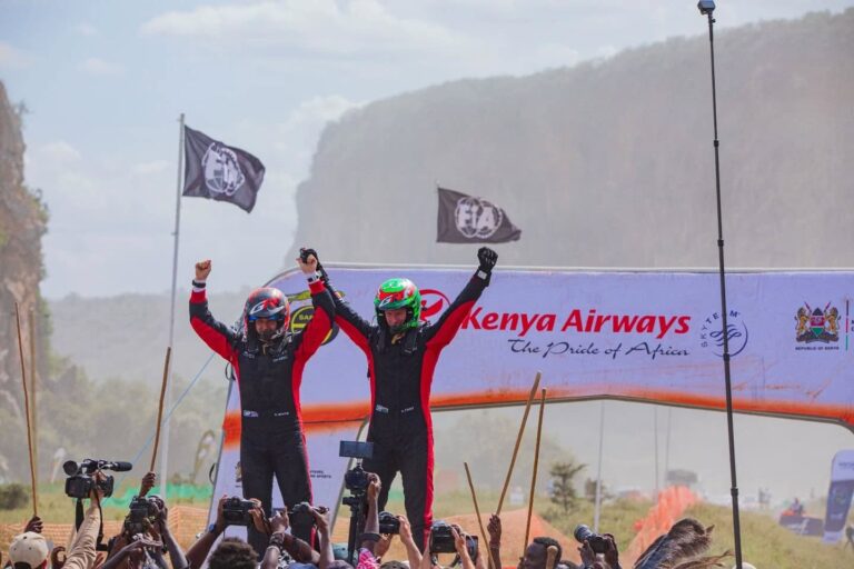 Two rally drivers celebrate at the finish line of the 2025 WRC Safari Rally in Kenya. Both are in full racing gear with raised arms in triumph, framed by a banner featuring “Kenya Airways” and “The Pride of Africa,” along with FIA and Republic of Kenya logos. Spectators and photographers gather to document the moment.