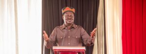 Raila Odinga speaking at a podium during a formal event, wearing traditional attire and gesturing with raised hands
