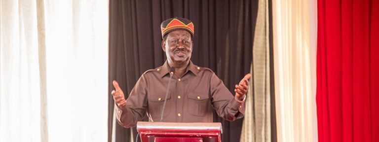 Raila Odinga speaking at a podium during a formal event, wearing traditional attire and gesturing with raised hands