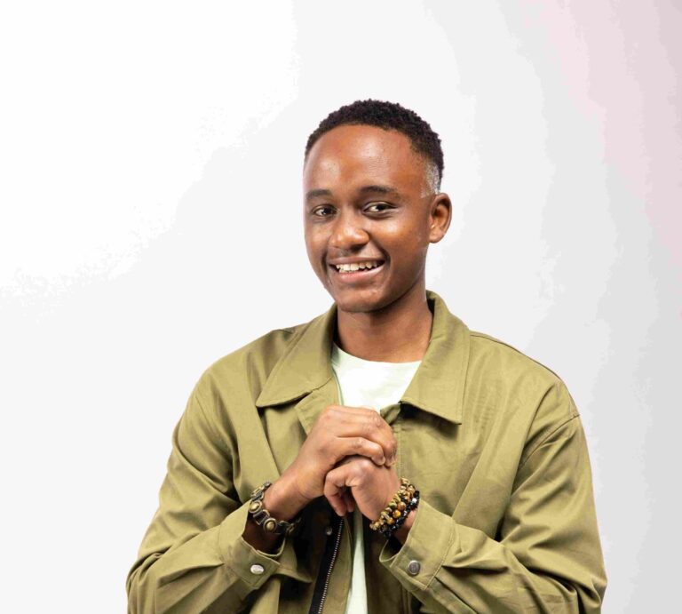 Okoyo Isaac stands smiling in a studio portrait, wearing layered green tones and beaded bracelets, hands clasped in front of his chest.