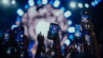xConcert crowd raising smartphones to record a live performance, with blue stage lighting and bright spotlights illuminating the scene from behind.