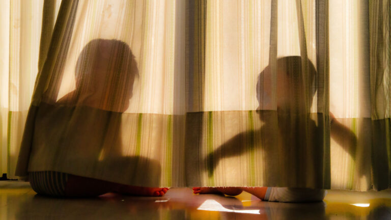Kenya’s Missing Children Crisis Demands Prevention Over Reaction Two children silhouette playfully sitting behind sheer curtains in sunlight.