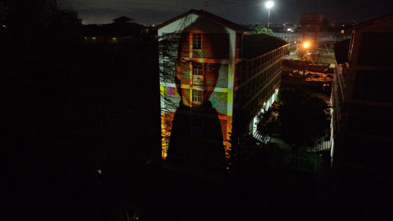 An aerial drone photograph taken at night in Nairobi shows a large projected portrait of a Kenyan person spanning the full facade of a multi-storey brick building, the face rendered in warm amber tones against the darkened surrounding neighbourhood.