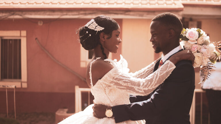 A joyful newlywed couple embraces outdoors on their wedding day, with the bride in a beaded white gown and the groom in a dark suit holding a bouquet of pink and white roses.
