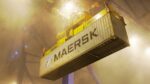 A Maersk shipping container lifted by a crane at a port terminal at night, surrounded by fog and floodlights.