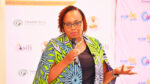 Ms. Winnie Ngumi speaking with a microphone in front of branded event backdrops featuring KAM (Kenya Association of Manufacturers) logos, Frametech Structures, and Asili Roofing Tiles.