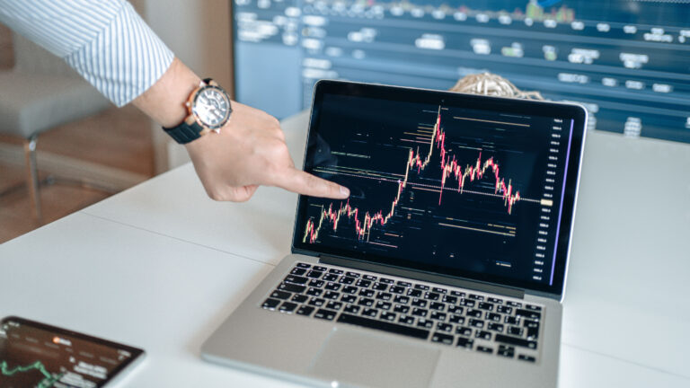 Trader points to live candlestick charts on a laptop screen displaying market trends