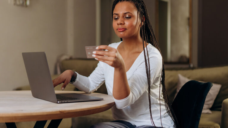 When Generosity in Love Becomes Financial Enabling A woman with braided hair sits at a table, holding a card up to read while her other hand rests on a laptop keyboard.