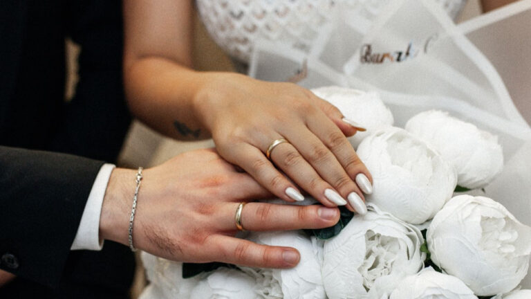 A romantic close-up of a bride and groom showcasing their wedding rings atop a white floral bouquet.