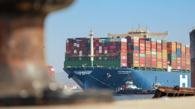 HMM Enters Mombasa With New India Container Service HMM container ship Algeciras stacked with containers, guided by two tugboats, seen past a blurred mooring bollard.