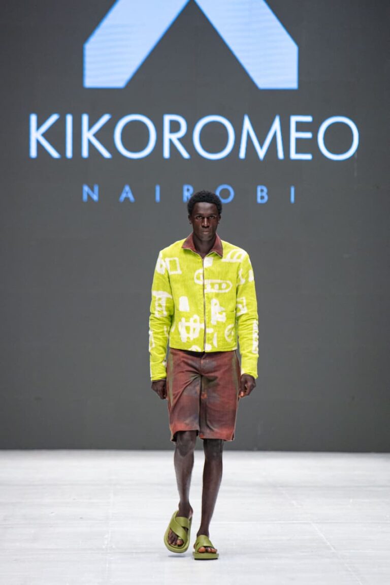 KikoRomeo Takes Kenyan Fashion To Moscow Fashion Week Model walks a runway in a lilac patterned long coat and matching trousers in front of a KikoRomeo Nairobi sign