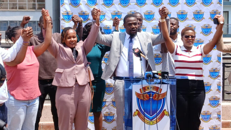 Doctors’ Union Gives Government Seven Days to End Nurses’ Strike KMPDU Secretary General Davji Atellah speaks at a podium during a press briefing, surrounded by union members raising joined hands in a show of solidarity.