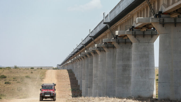 A red four wheel drive vehicle drives along a dirt road beside a long elevated railway viaduct. Rows of concrete pillars carry the track across dry grassland under a clear sky.