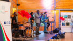 Four musicians on a stage at Shamba Events: a drummer, a bass guitarist, a trumpeter and a saxophonist. An Embassy of Italy Nairobi banner stands at left and a display of Italian gin labels hangs at right.