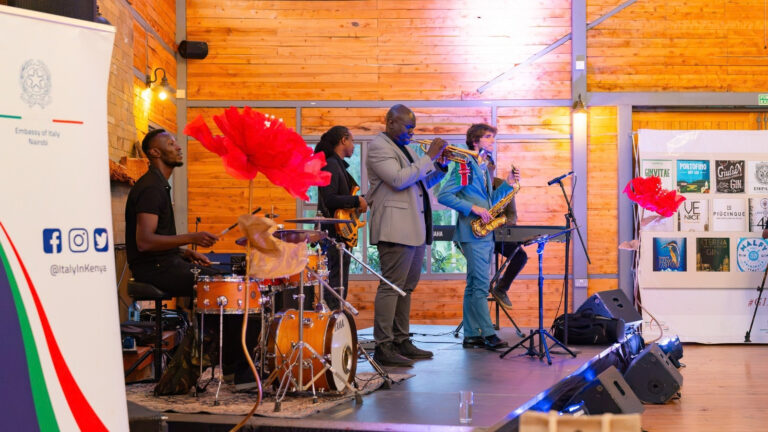 Italy Brings Gin, Jazz and Cars to Nairobi Four musicians on a stage at Shamba Events: a drummer, a bass guitarist, a trumpeter and a saxophonist. An Embassy of Italy Nairobi banner stands at left and a display of Italian gin labels hangs at right.