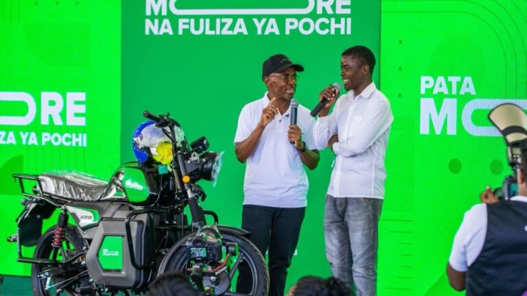 Safaricom Group CEO Peter Ndegwa and a young man speak into microphones on stage beside a green motorbike, under a “Pata More na Fuliza Ya Pochi” banner.