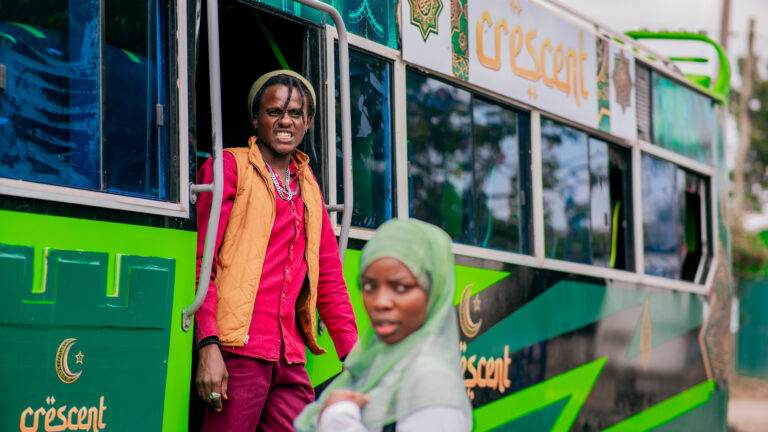 Crescent on the Road Film Premieres in Kilimani A green matatu with gold “Crescent” lettering. A man in a beanie, red shirt and orange vest stands in the open doorway, and a woman in a pale green hijab is in the foreground.